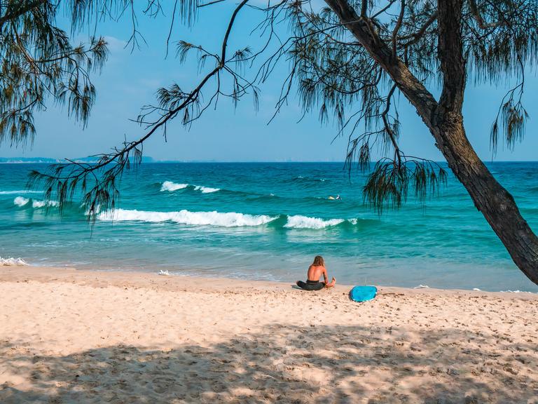 Ein Surfer sitzt mit seinem Surfbrett am Rainbow Bay Beach in Australien.