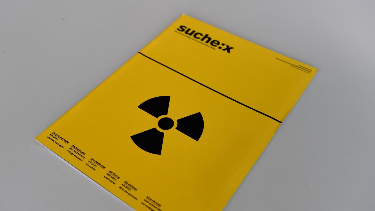 Eine Broschüre mit der Aufschrift "Suche:X" und einem Strahlenwarnzeichen liegt bei einer Pressekonferenz auf einem Tisch. 