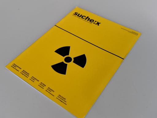 Eine Broschüre mit der Aufschrift "Suche:X" und einem Strahlenwarnzeichen liegt bei einer Pressekonferenz auf einem Tisch. 