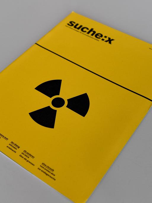 Eine Broschüre mit der Aufschrift "Suche:X" und einem Strahlenwarnzeichen liegt bei einer Pressekonferenz auf einem Tisch. 