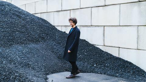 Ein junger Mann mit Brille und lockigen Haaren steht vor einer Wand am Rande einer Koks-Halde
