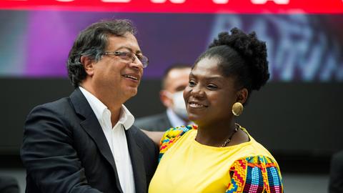 Ein Herr mit Jacket und weißem Hemd und eine Afrokolumbianerin mit gelbem Kleid nehmen sich lächelnd in den Arm. Ein Herr mit Jacket und weißem Hemd und eine Afrokolumbianerin mit gelbem Kleid nehmen sich lächelnd in den Arm.