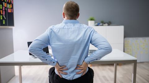 Ein Mann sitzt am Schreibtisch und hält beide Hände gegen den unteren Rücken. (Symbolfoto)