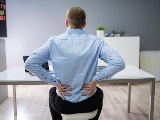 Ein Mann sitzt am Schreibtisch und hält beide Hände gegen den unteren Rücken. (Symbolfoto)