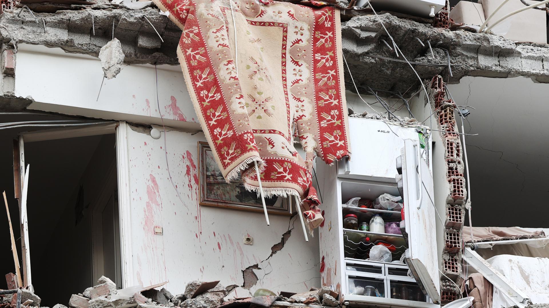 Ein vom Erdbeben zerstörtes Gebäude. Am Haus steht ein Regal, auf dem die Habe der Bewohner zu erkennen ist, aus dem oberen, unter Trümmern liegenden Stockwerk hängt ein bunter Teppisch herab.