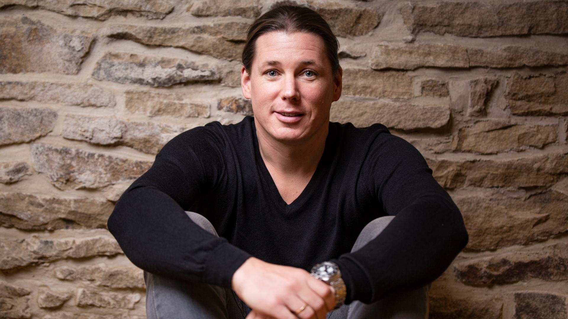 Michele Ufer trägt einen dunklen Pullover und eine graue Jeans. Er sitzt auf dem Boden und schaut freundlich in die Kamera. Im Hintergrund ist eine gemauerte Natursteinwand zu sehen. Michele Ufer trägt einen dunklen Pullover und eine graue Jeans. Er sitzt auf dem Boden und schaut freundlich in die Kamera. Im Hintergrund ist eine gemauerte Natursteinwand zu sehen.