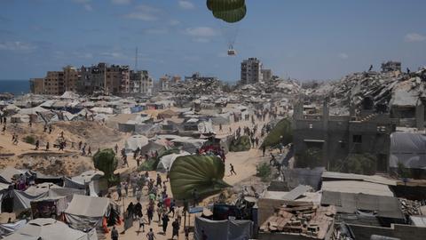 Palästinenser sammeln Hilfsgüter ein, die mit Fallschirmen über Gaza-Stadt abgeworfen wurden. Im Hintergrund Zelte und zerstörte Gebäude