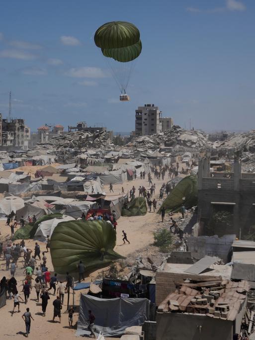 Palästinenser sammeln Hilfsgüter ein, die am 7. August 2025 mit Fallschirmen über Gaza-Stadt im nördlichen Gazastreifen abgeworfen werden. Im Hintergrund Fallschirme, Zelte, zerstörte Gebäude und das Mittelmeer.
