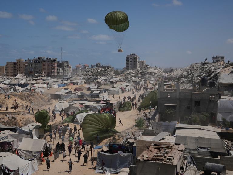 Palästinenser sammeln Hilfsgüter ein, die am 7. August 2025 mit Fallschirmen über Gaza-Stadt im nördlichen Gazastreifen abgeworfen werden. Im Hintergrund Fallschirme, Zelte, zerstörte Gebäude und das Mittelmeer.