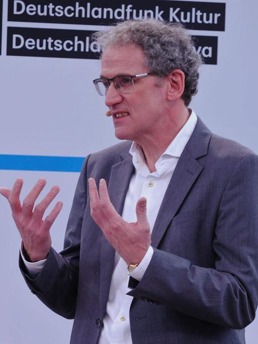 Dirk Oschmann in weissem Hemd und grauer Jacke spricht vor einer weissen Wand mit Deutschlandradio Logos im Hintergrund.