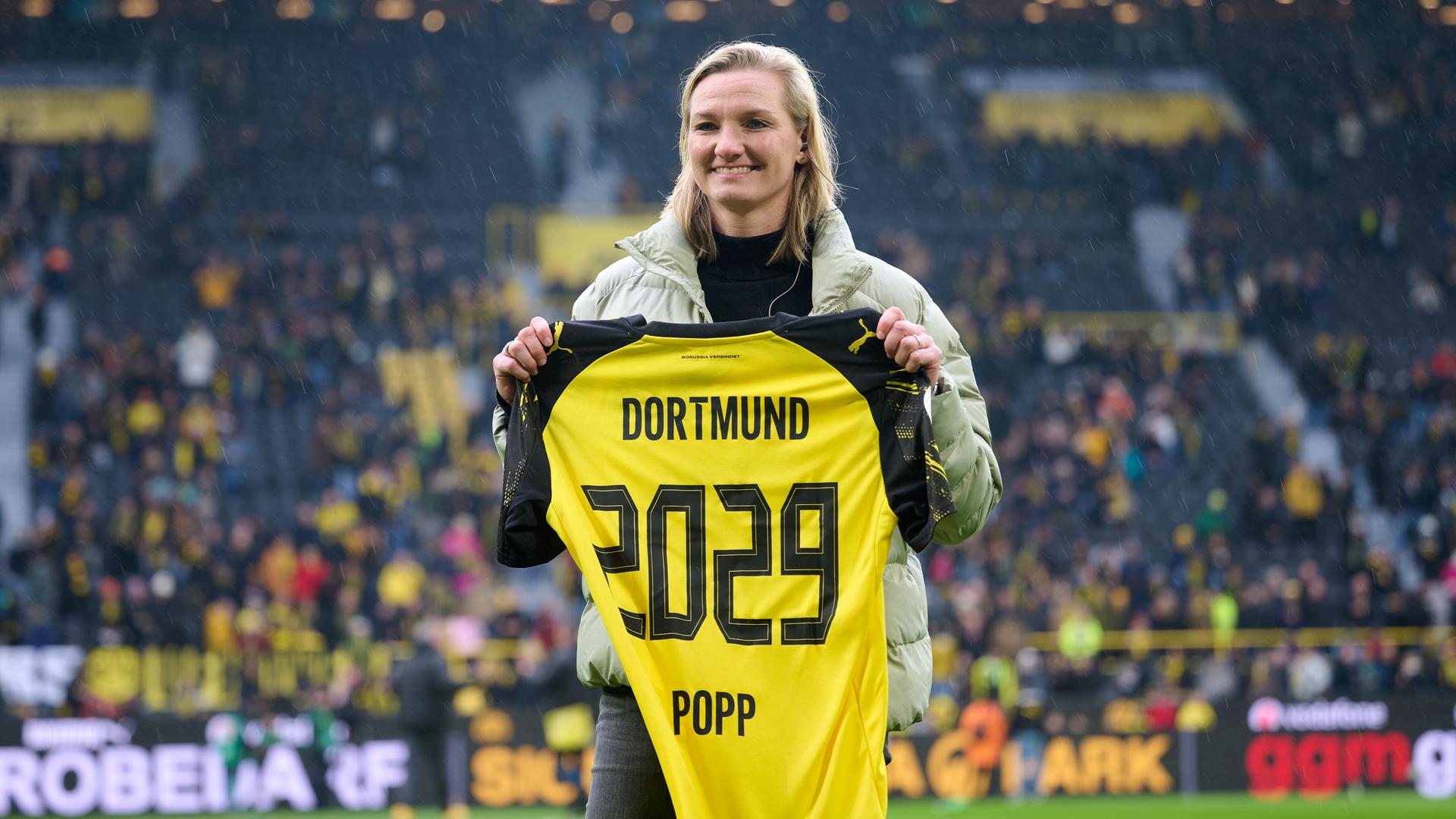 Alexandra Popp steht mit einem Trikot ihres neuen Vereins Borussia Dortmund mit der Nummer 2029 auf dem Rasen des Signal-Iduna-Parks und lacht. Alexandra Popp steht mit einem Trikot ihres neuen Vereins Borussia Dortmund mit der Nummer 2029 auf dem Rasen des Signal-Iduna-Parks und lacht.