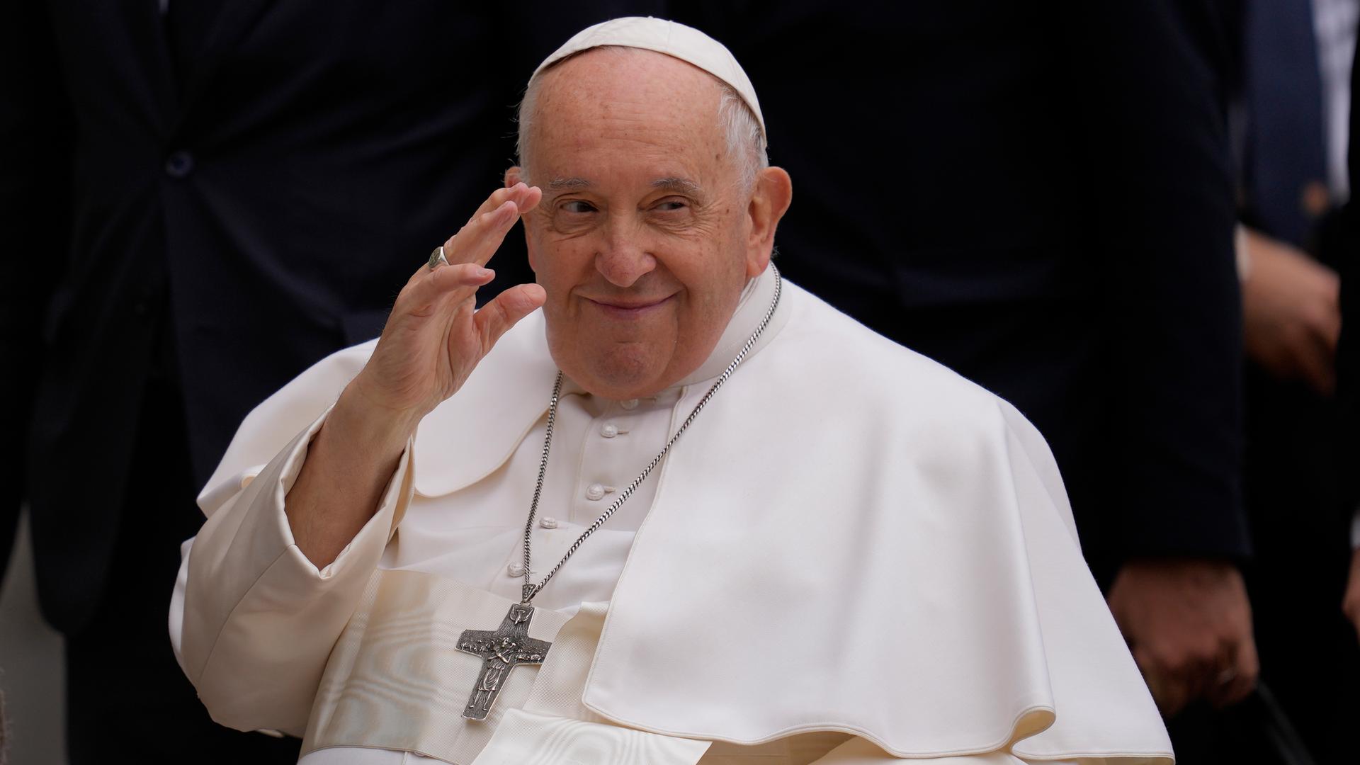 Der Papst sitzt im Rollstuhl und winkt. Er trägt ein weißes Gewand und ein großes Kreuz um den Hals.