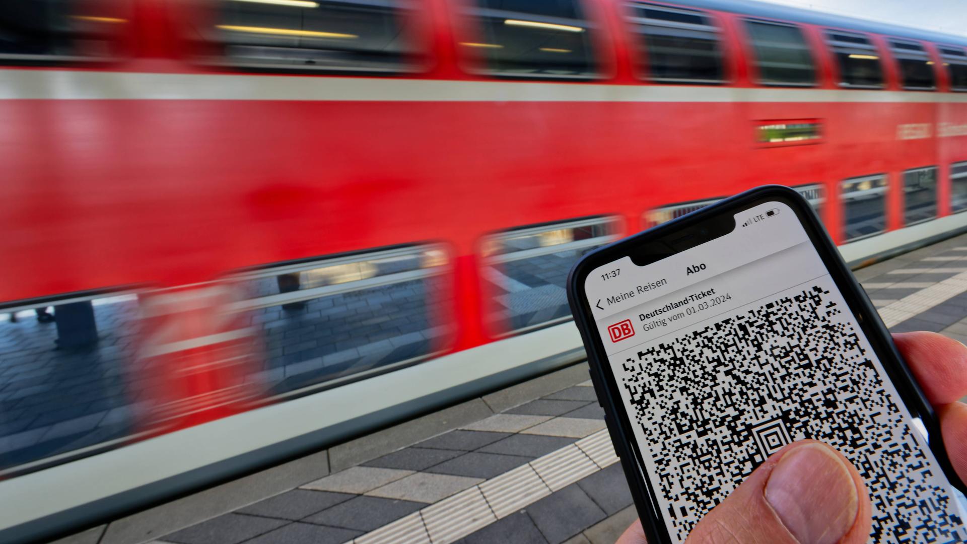 Das Deutschlandticket auf einem Smartphone an einem Bahnsteig. Im Hintergrund fährt ein Zug vorbei.