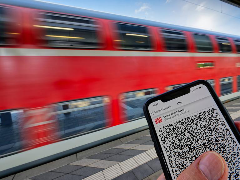 Das Deutschlandticket auf einem Smartphone an einem Bahnsteig. Im Hintergrund fährt ein Zug vorbei