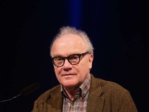 Ein Mann mit kurzem grauen Haar und schwarz umrandeter Brille spricht vor dunklem Hintergrund in ein Mikrofon, im Rahmen des Kölner Literaturfestivals Lit.Cologne im Jahr 2022.