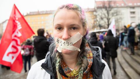 Eine junge Frau bei einer Demo. Sie hat mit Klebeband ein Kreuz über den Mund geklebt, das es ihr unmöglich macht zu sprechen. Auf dem Klebeband steht "Paragraf 219a"