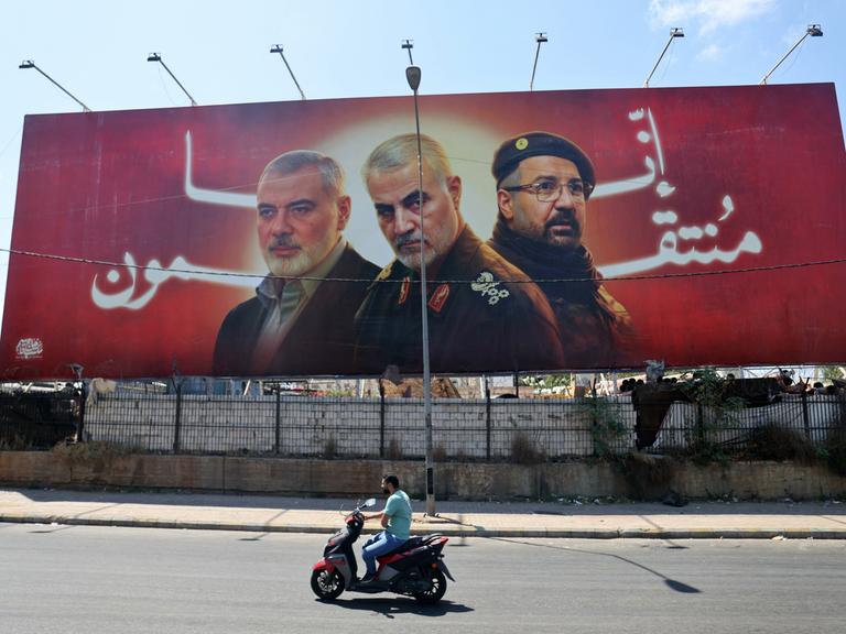 An einer Straße steht ein Banner mit Porträts der getöteten Milizenführer Ismail Haniyeh, Qasem Soleimani und Fuad Shukr.