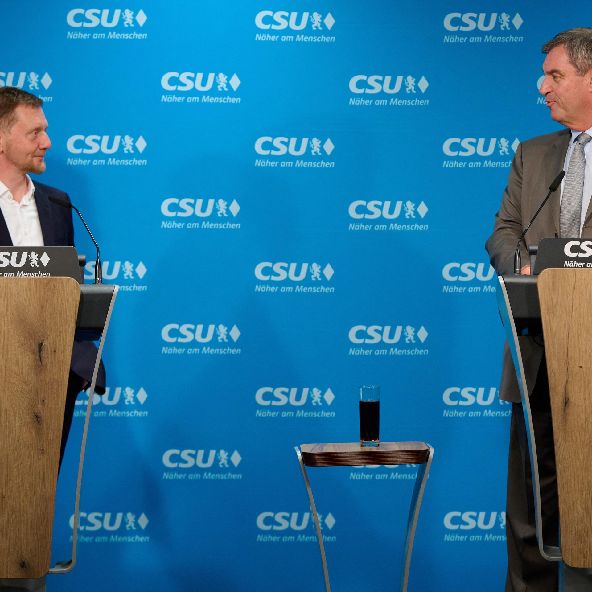 Markus Söder und Michael Kretschmer stehen an zwei nebeneinander stehenden Stehpulten und sehen sich an. An der Wand hinter ihnen prangt das Logo der CSU.