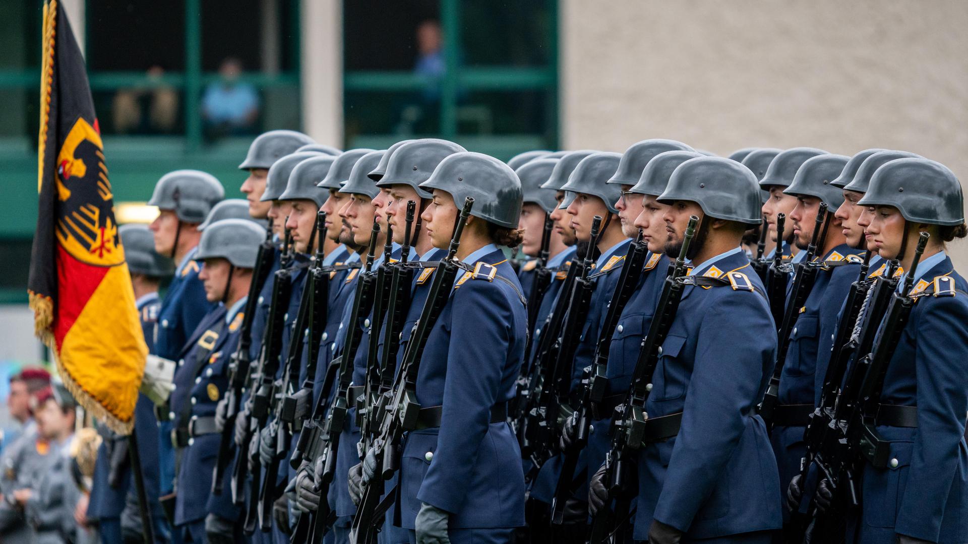 Appell zur Verabschiedung des 124. Offizierslehrgangs der Luftwaffe der Bundeswehr in der Kaserne in Fürstenfeldbruck. Soldaten in blauen Uniformen und Helmen stehen in Formation mit Gewehren. Neben ihnen eine Fahne mit dem Symbol des Bundesadlers.