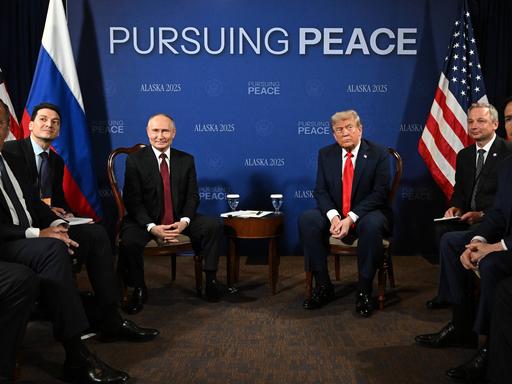 Sechs Männer sitzen nebeneinander auf Stühlen, in der Mitte der russische Staatschef Putin und US-Präsident Trump.