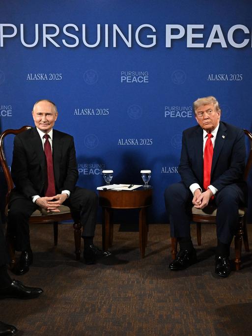 Sechs Männer sitzen nebeneinander auf Stühlen, in der Mitte der russische Staatschef Putin und US-Präsident Trump.