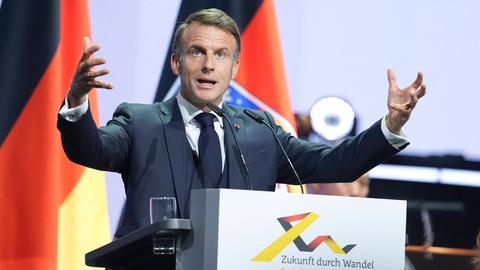 Ein Mann im Anzug steht am Rednerpult. Er hebt die Arme.