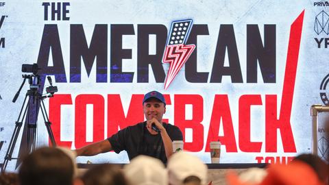 Charlie Kirk im T-Shirt und mit Basecap spricht vor einem Plakat mit der Aufschrift "The American Comeback" in ein Mikrofon