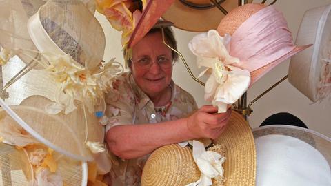 Firmenchefin Heide Steyer, eine ältere Dame in kurzärmeliger Bluse, ist umringt von Hüten mit großen Kunstblumen in verschiedenen Größen und Formen. Ihre Manufaktur im sächsischen Wallroda stellt die Stoffblumen her.