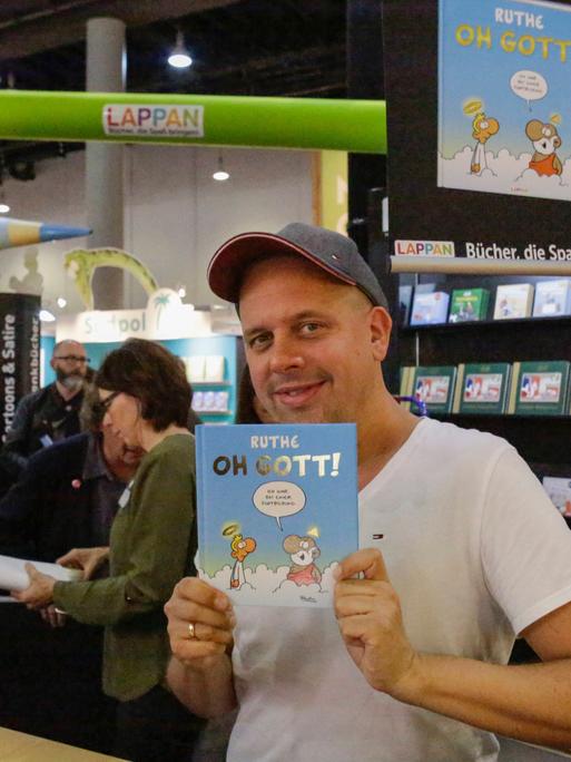Der Karikaturist Ralph Ruthe hält auf der Frankfurter Buchmesse 2019 sein Cartoon-Buch mit dem Titel "Oh Gott" in die Kamera und lächelt. 

