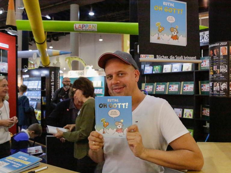Der Karikaturist Ralph Ruthe hält auf der Frankfurter Buchmesse 2019 sein Cartoon-Buch mit dem Titel "Oh Gott" in die Kamera und lächelt. 

