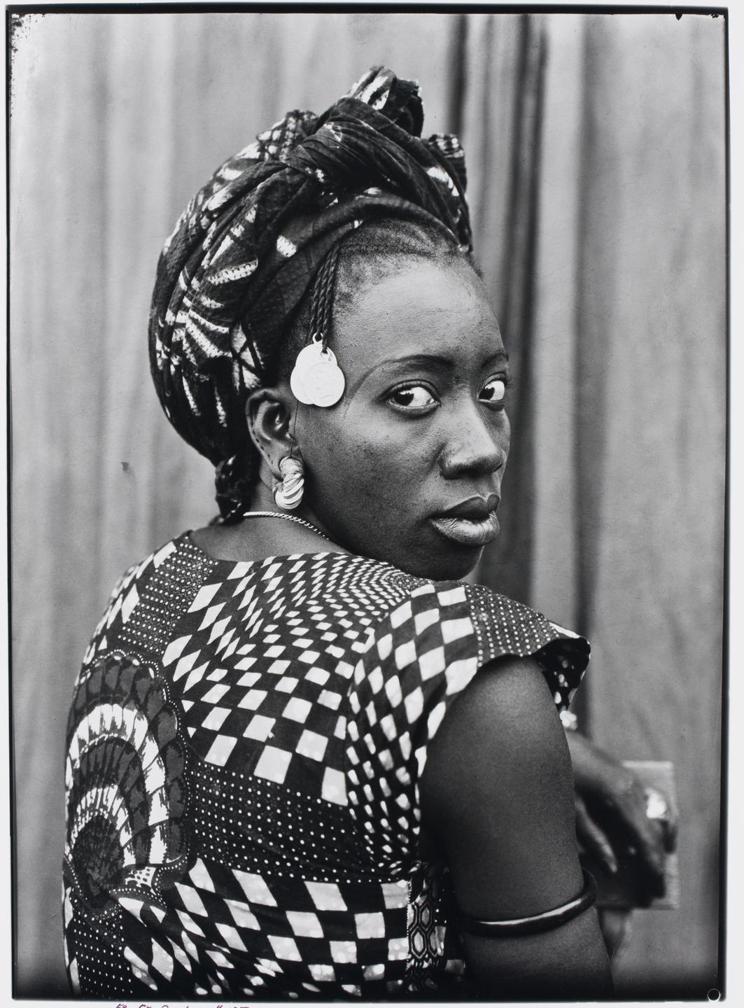 Schwarzweißfotografie einer Frau aus Mali. Sie trägt ein kariertes Kleid und ein passendes Kopftuch und schaut den Betrachter über die Schulter an.