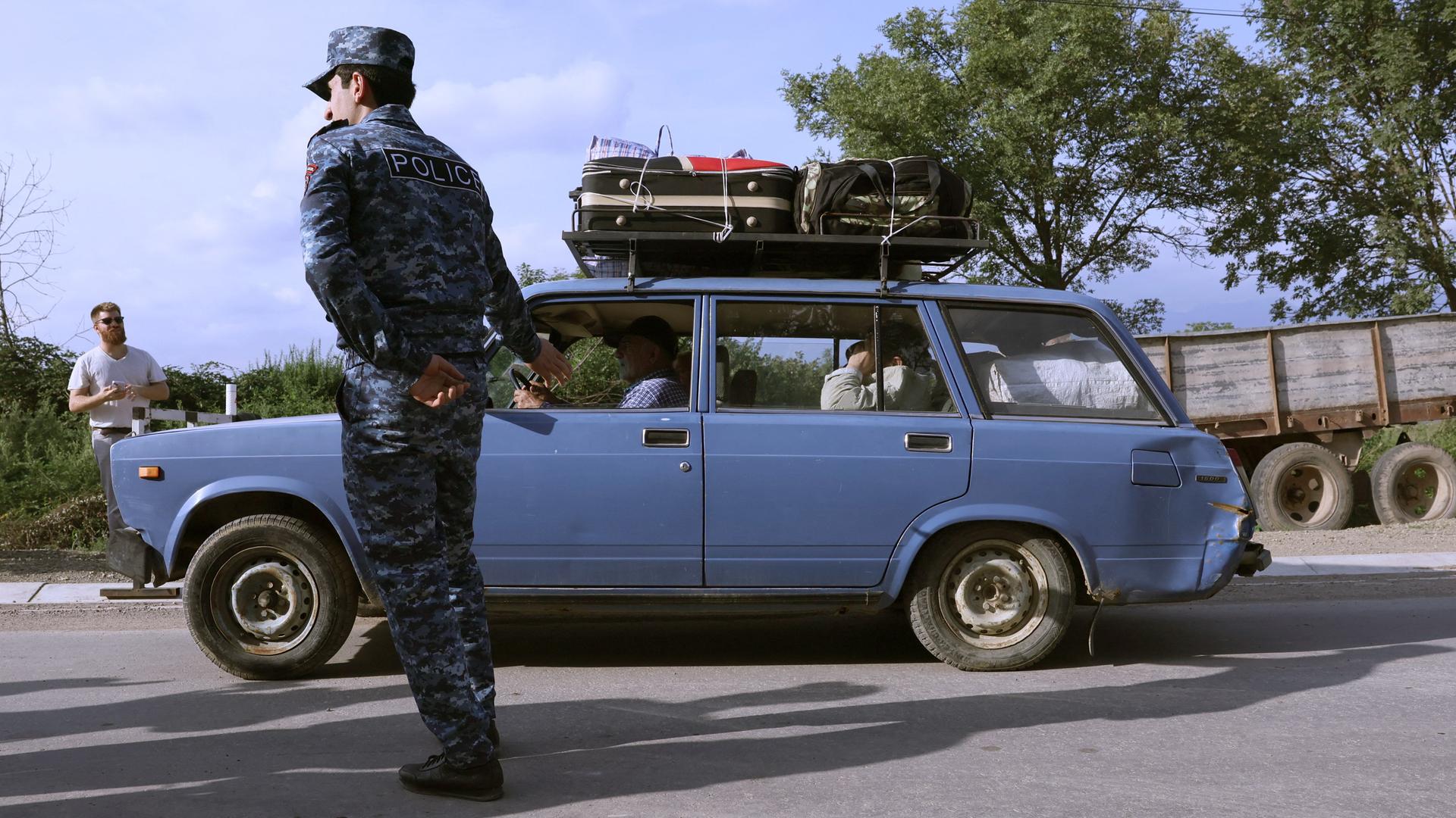 Ein Polizist winkt ein blaues Auto durch, auf dessen Dach mehrere Koffer montiert sind.