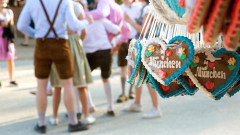 Lebkuchenherzen mit der Aufschrift "I love München" hängen an einem Stand auf der Theresienwiese. Im Hintergrund stehen feiernde Menschen in Lederhosen und Dirndl.