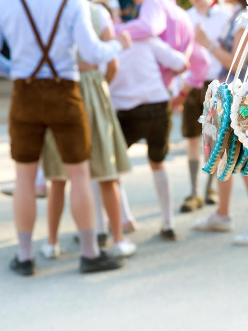 Lebkuchenherzen mit der Aufschrift "I love München" hängen an einem Stand auf der Theresienwiese. Im Hintergrund stehen feiernde Menschen in Lederhosen und Dirndl.