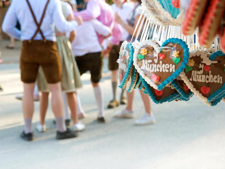 Lebkuchenherzen mit der Aufschrift "I love München" hängen an einem Stand auf der Theresienwiese. Im Hintergrund stehen feiernde Menschen in Lederhosen und Dirndl.