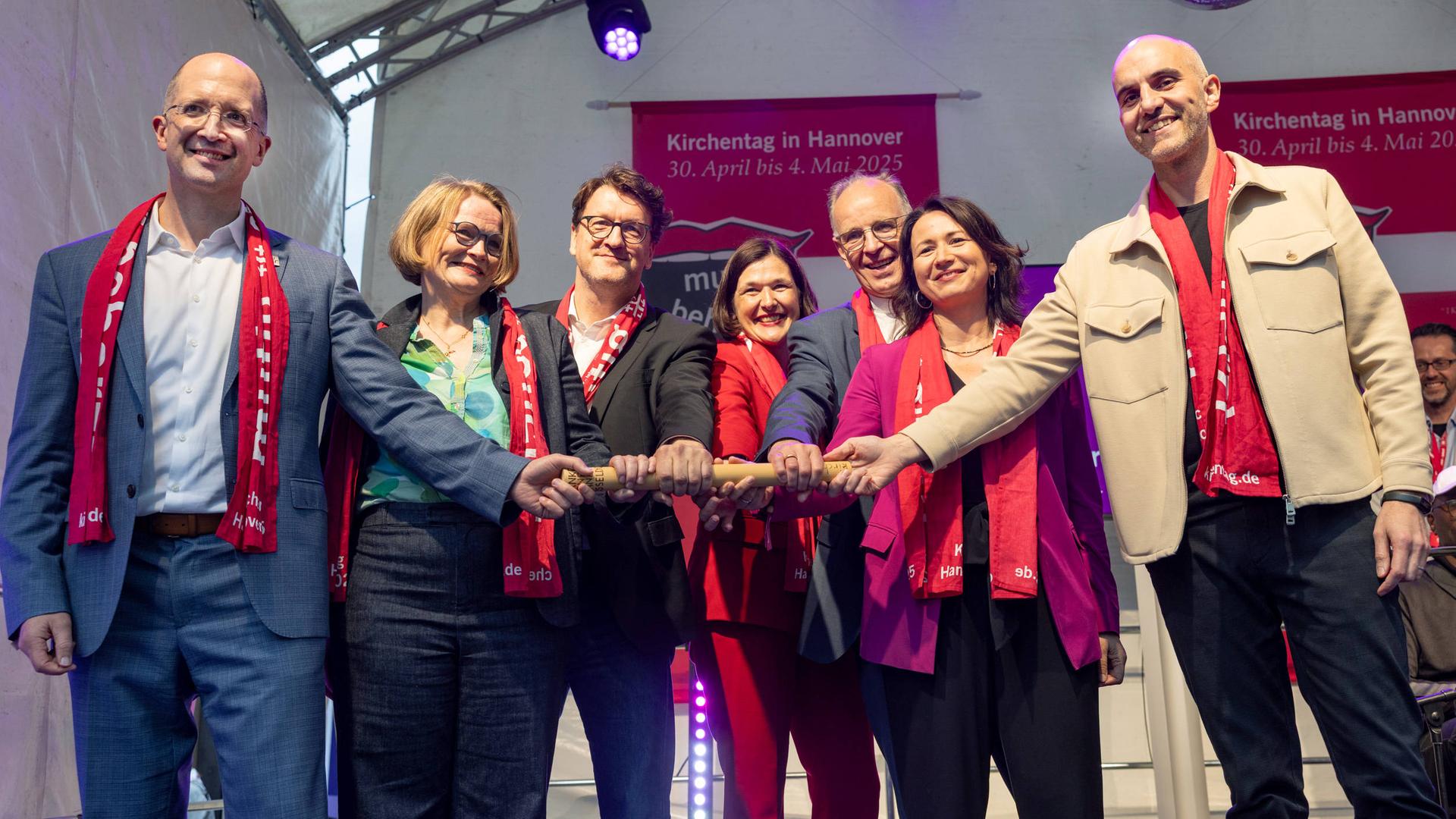 Das Foto zeigt eine Gruppe mit insgesamt sieben Menschen. In ihrer Mitte ist ein Staffelstab zu sehen, den alle gemeinsam festhalten. Das Foto zeigt eine Gruppe mit insgesamt sieben Menschen. In ihrer Mitte ist ein Staffelstab zu sehen, den alle gemeinsam festhalten.