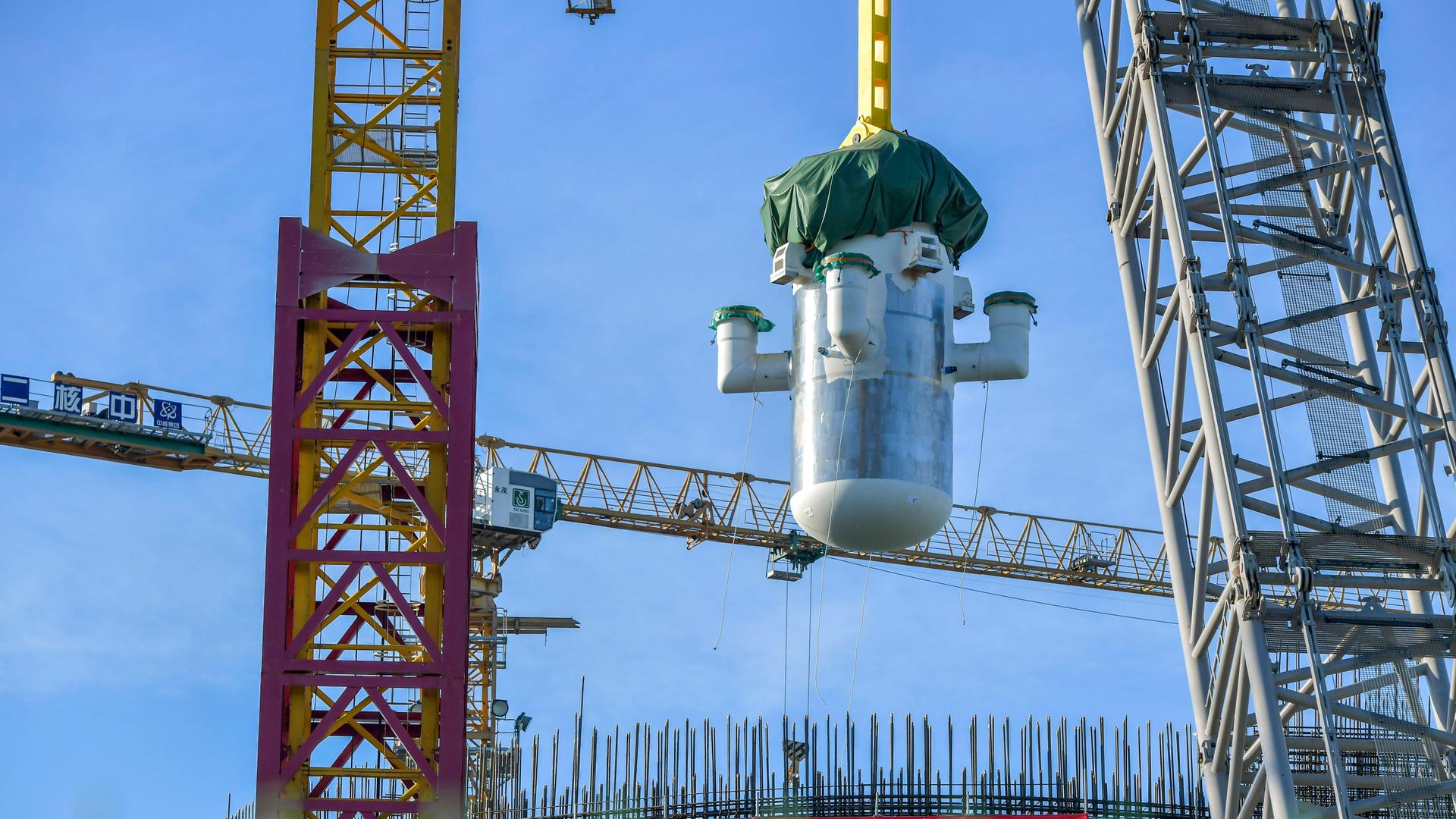 Per Kran schwebt ein großes zylindrisches Bauteil eines chinesischen Mini-AKWs über einer Baustelle mit Gerüsten und Stahlträgern vor blauem Himmel.