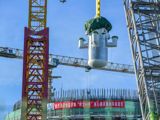Per Kran schwebt ein großes zylindrisches Bauteil eines chinesischen Mini-AKWs über einer Baustelle mit Gerüsten und Stahlträgern vor blauem Himmel.
