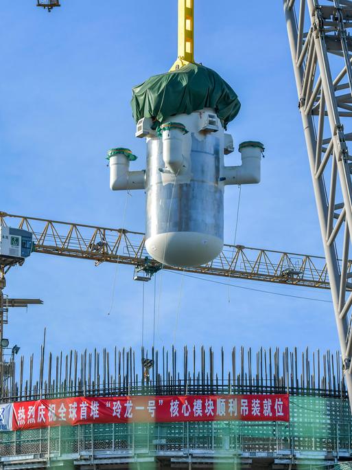 Per Kran schwebt ein großes zylindrisches Bauteil eines chinesischen Mini-AKWs über einer Baustelle mit Gerüsten und Stahlträgern vor blauem Himmel.