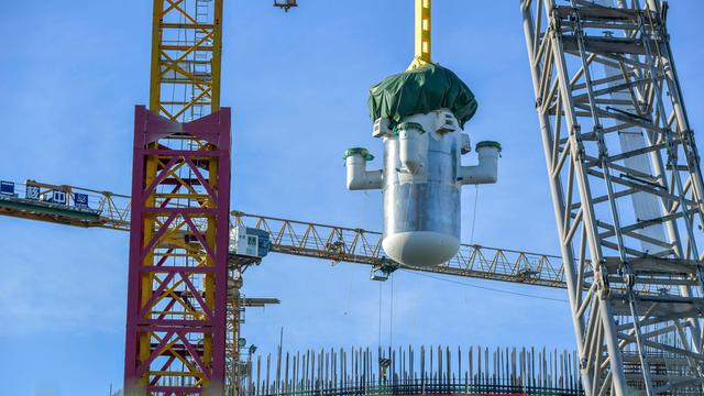 Per Kran schwebt ein großes zylindrisches Bauteil eines chinesischen Mini-AKWs über einer Baustelle mit Gerüsten und Stahlträgern vor blauem Himmel.