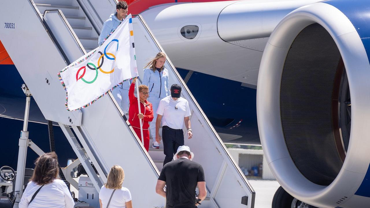 Zu sehen ist, wie die Bürgermeisterin von Los Angeles Karen Bass aus dem Flugzeug steigt und die Olympische Flagge hoch hält.