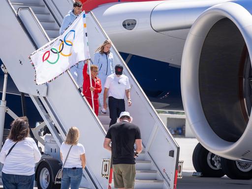 Zu sehen ist, wie die Bürgermeisterin von Los Angeles Karen Bass aus dem Flugzeug steigt und die Olympische Flagge hoch hält.