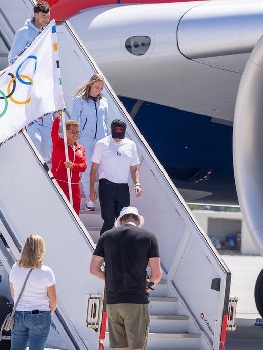 Zu sehen ist, wie die Bürgermeisterin von Los Angeles Karen Bass aus dem Flugzeug steigt und die Olympische Flagge hoch hält.