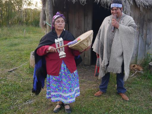 Eine Frau in traditioneller Kleidung und großem Silberschmuck am Hals spielt eine Trommel, während ein mann, ebenfalls mit traditionellem Umhang, ein Blasinstrument spielt.