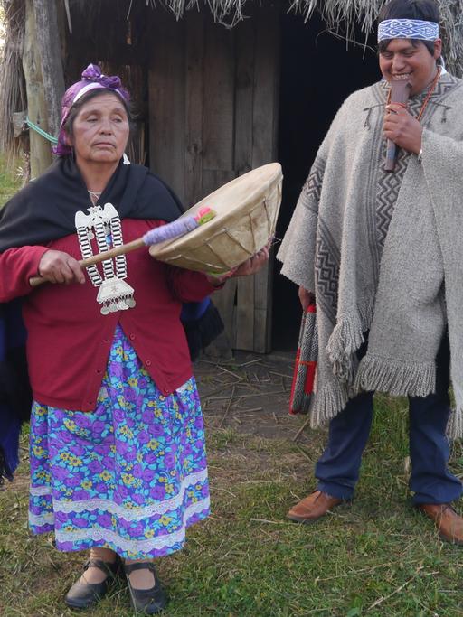 Eine Frau in traditioneller Kleidung und großem Silberschmuck am Hals spielt eine Trommel, während ein mann, ebenfalls mit traditionellem Umhang, ein Blasinstrument spielt.