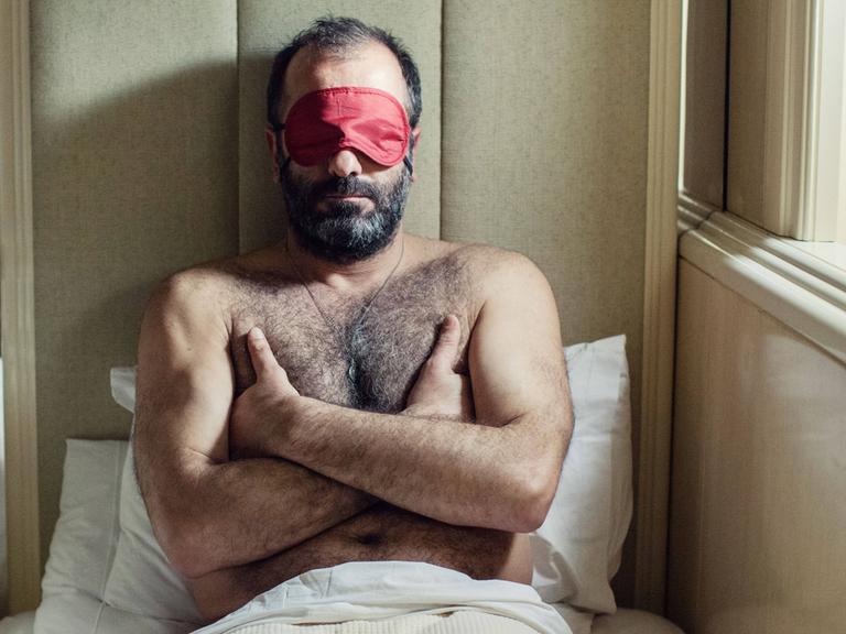 Ein Mann mit ausgeprägter Brustbehaarung sitzt im Bett und trägt eine Schlafmaske.