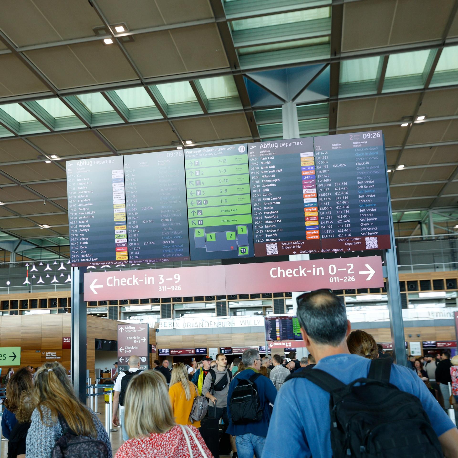 Menschen stehen in einer Halle des Berliner Flughafens BER und schauen auf eine Anzeigetafel.
