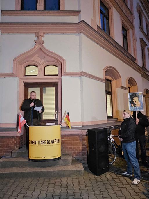 Ein Redner der Identitären Bewegung spricht vor dem "Zentrum Chemnitzer", einem weiß-roten Eckgebäude. Um ihn herum stehen Pressevertreter mit Kameras und Mikrofonen.
