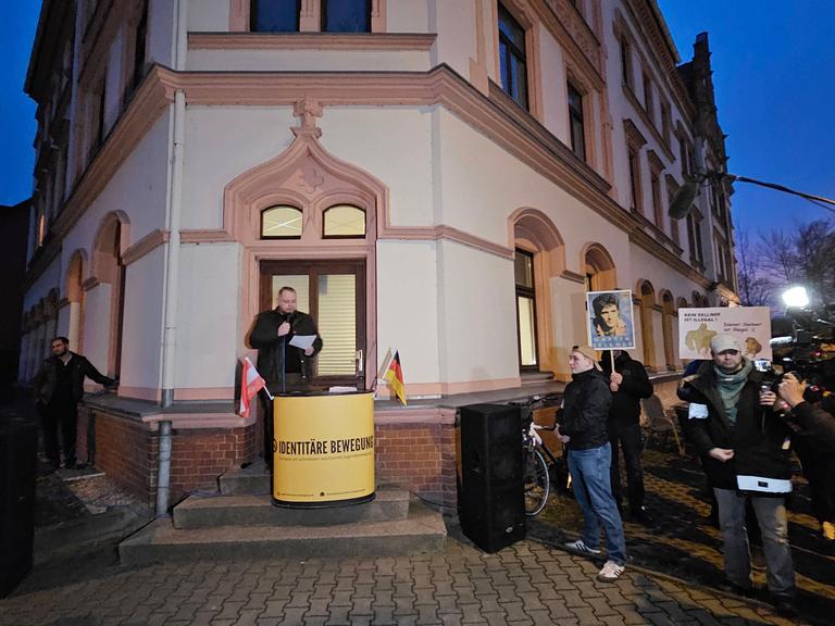 Ein Redner der Identitären Bewegung spricht vor dem "Zentrum Chemnitzer", einem weiß-roten Eckgebäude. Um ihn herum stehen Pressevertreter mit Kameras und Mikrofonen.