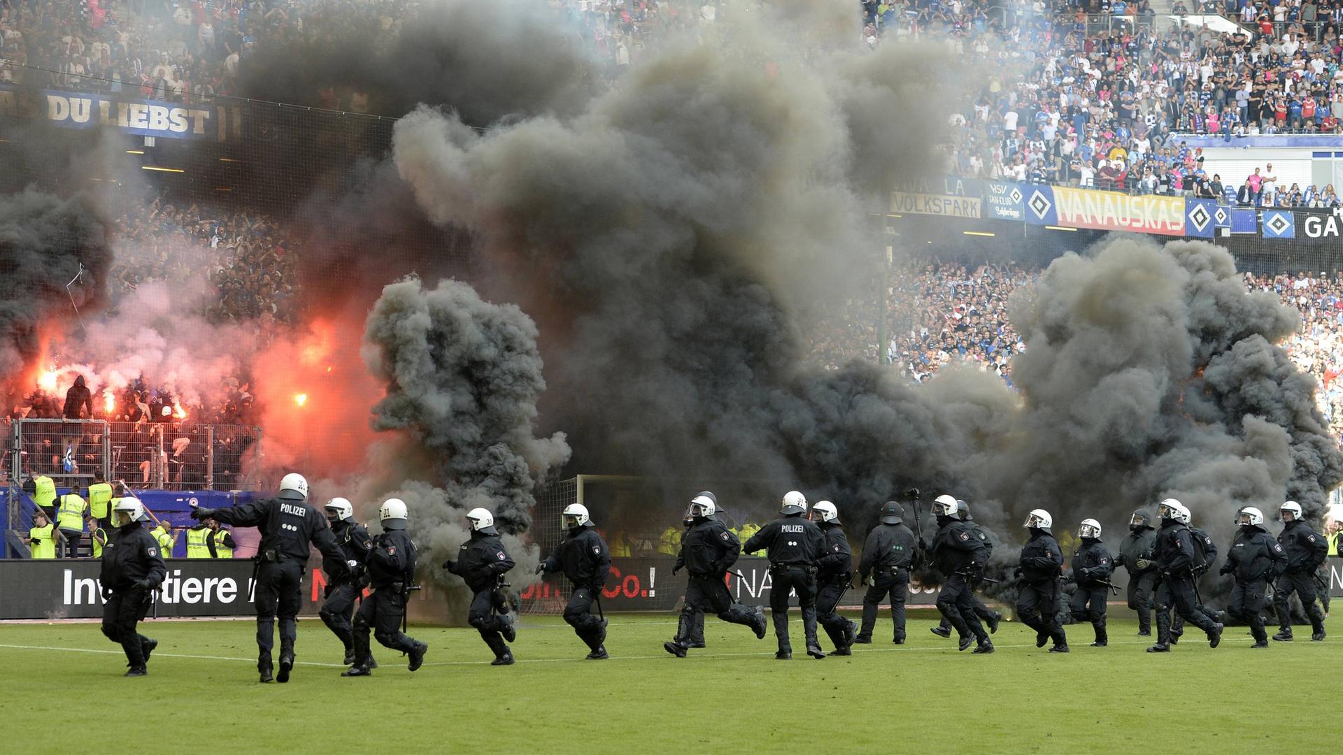 Polizisten rennen in Kampfmontur auf einem Fußballfeld. Die Zuschauerränge werden von Flammen und Rauch verdeckt.